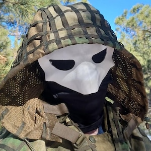 Op de afbeelding: Een camouflagekap met een wit masker met zwarte ooggaten en een zwarte gezichtsbedekking. De kap heeft een netontwerp en maakt deel uit van een outfit in militaire stijl.