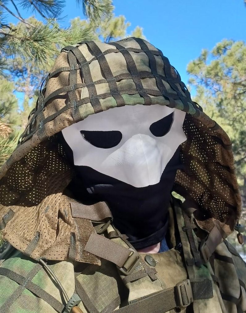 Op de afbeelding: Een camouflagekap met een wit masker met zwarte ooggaten en een zwarte gezichtsbedekking. De kap heeft een netontwerp en maakt deel uit van een outfit in militaire stijl.
