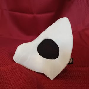 Op de afbeelding: Een wit masker met een zwarte oogopening. De mask is geplaatst tegen een rode achtergrond. Het masker is gemaakt van een glad, stevig materiaal en heeft een gebogen vorm, wat suggereert dat het is ontworpen om op het gezicht te passen.