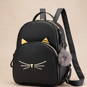 Może przedstawiać: Czarny plecak z motywem kociej twarzy. Plecak posiada złote kocie uszy, złoty zamek błyskawiczny i szary brelok z pomponem. Twarz kota jest haftowana złotą nicią.