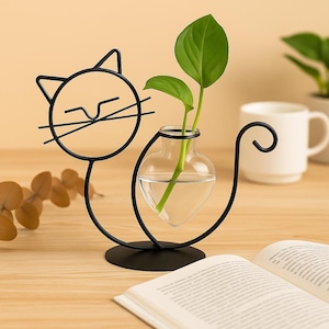 Könnte beinhalten: Eine schwarze Metallvase in Katzenform, die eine klare Glasvase mit Wasser und grünen Pflanzenstängeln hält. Das Gesicht der Katze ist ein einfacher Umriss, und der Schwanz krümmt sich nach oben. Die Vase steht auf einem runden Sockel.