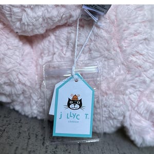 May include: A soft, pink plush toy with a tag attached. The tag has a black cat wearing a crown, the text "j llyc T. LONDON", and a teal border. The toy has a fluffy, textured appearance.