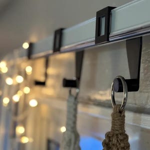 May include: Black metal curtain rod hooks attached to a white curtain rod. The hooks are holding macrame plant hangers. A string of warm white fairy lights is in the background.