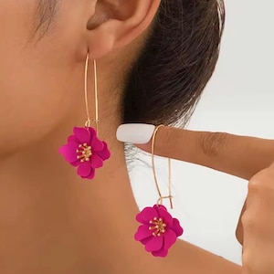 Könnte beinhalten: Hängeohrringe mit leuchtend pinkfarbenen Blumenanhängern mit goldfarbenen Zentren und langen, dünnen goldfarbenen Haken. Die Ohrringe werden vor einem neutralen Hintergrund gezeigt, der die lebendige Farbe des floralen Designs hervorhebt.