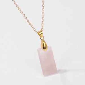 Könnte beinhalten: Halskette mit Anhänger aus Rosenquarz und goldfarbener Kette. Der rechteckige Anhänger hat eine zarte rosa Farbe. Die Halskette wird vor einem weißen Hintergrund präsentiert.