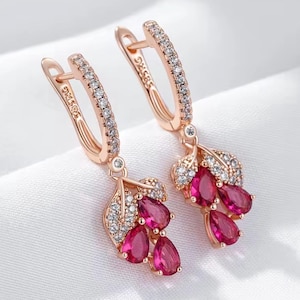 Könnte beinhalten: Ohrringe aus Roségold mit Blattdesign. Jeder Ohrring hat einen gebogenen Bügel mit kleinen, klaren Steinen und einen hängenden Cluster aus drei tropfenförmigen, roten Edelsteinen. Auf weißem Hintergrund.