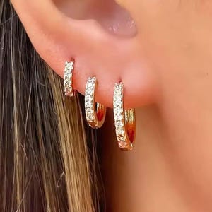 Könnte beinhalten: Drei goldene Creolen, jeweils besetzt mit kleinen, klaren Edelsteinen. Die Ohrringe sind unterschiedlich groß, der kleinste im oberen Piercing und der größte im unteren. Die Ohrringe werden an einem Ohr getragen.