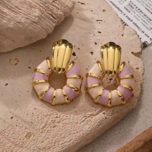 Könnte beinhalten: Ein Paar goldfarbene Ohrringe mit einzigartigem Design. Jeder Ohrring hat ein goldfarbenes Oberteil mit fächerförmiger Form, verbunden mit einem kreisförmigen Anhänger mit abwechselnden Abschnitten in Hellrosa, Weiß und Gold. Auf neutralem Hintergrund.