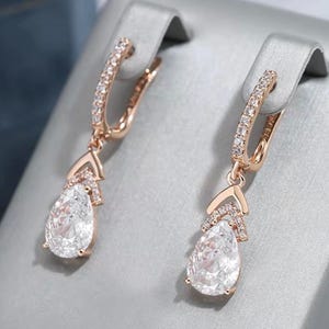 Könnte beinhalten: Ohrringe in Roségold mit einem tropfenförmigen, klaren Edelstein. Die Ohrringe haben einen Klappverschluss und sind mit kleinen, funkelnden Steinen am oberen Teil und am Verbindungspunkt des Tropfens verziert.