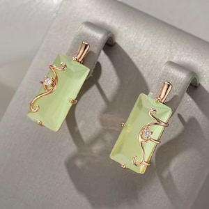 May include: A pair of rectangular earrings featuring a pale green gemstone. The earrings have a gold-coloured frame with a decorative swirl and a small, clear gemstone accent. The earrings are displayed on a grey surface.