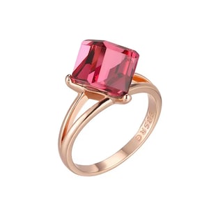 May include: A rose gold ring featuring a large, rectangular, faceted pink gemstone. The band splits into two graceful curves as it rises to meet the gemstone setting. The ring is set against a white background.