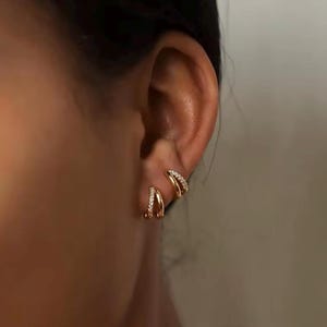 Op de afbeelding: Goudkleurige dubbele oorringen met kleine, sprankelende steentjes. De oorbellen worden op het oor gedragen en benadrukken hun delicate ontwerp en hoe ze het licht vangen. De sieraden voegen een vleugje elegantie toe.