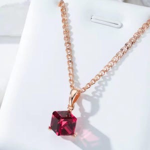 Könnte beinhalten: Halskette aus Roségold mit einem quadratischen, tiefroten Edelsteinanhänger. Der Anhänger hängt an einer zarten Kette. Die Halskette wird vor einem weißen Hintergrund gezeigt, um das Design und die Farbe des Schmucks hervorzuheben.
