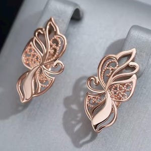 Könnte beinhalten: Ohrringe in Roségold-Optik mit Schmetterlingsmotiv, filigranem Muster und polierter Oberfläche. Die Ohrringe haben ein zartes Design mit geschwungenen Linien und einem dezenten Schimmer.