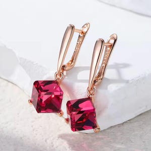 Könnte beinhalten: Ein Paar roségoldene Ohrringe mit quadratischen, facettierten, rubinroten Edelsteinen. Die Ohrringe haben einen Klappverschluss. Die Edelsteine haben eine tiefe, satte rote Farbe und fangen das Licht ein.
