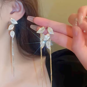 Könnte beinhalten: Elegante Ohrringe in Blattform mit funkelnden, klaren Kristallen. Die Ohrringe haben oben eine Ansammlung von drei blattförmigen Designs mit langen, zarten Metallsträngen, die darunter herabhängen. Die Ohrringe sind so konzipiert, dass sie baumeln und das Licht einfangen.