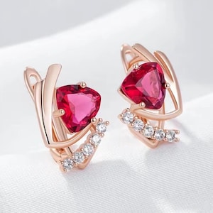 Könnte beinhalten: Ohrringe aus Roségold mit herzförmigen roten Edelsteinen und einer Reihe kleiner, klarer Kristalle. Die Ohrringe haben einen Klappverschluss. Auf weißem Hintergrund.