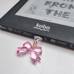 May include: A pink, faceted bow charm with a gold-coloured ring attached to a clear plastic clip. The charm is next to a black Kobo e-reader, displaying text that reads "FROM THE". The e-reader shows "51% read".
