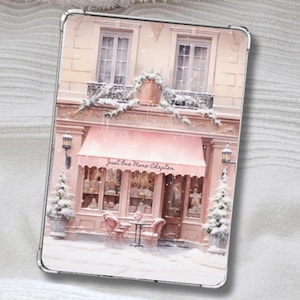 May include: A rectangular tablet case featuring a printed illustration of a pink storefront in winter. The shop has a pink awning with the text Just One More Chapter, two small tables with chairs, snow-covered trees, and a building facade with windows.