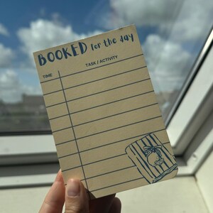 May include: A rectangular beige notepad with dark blue text and lines. The header reads BOOKED for the day, with columns for TIME and TASK / ACTIVITY. The bottom right corner features a line art illustration of a lemon on a striped towel.