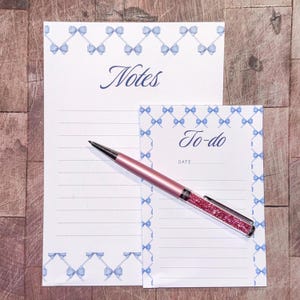 May include: A stationery set comprising a notepad and a to-do list pad, both with a white background and a light blue bow pattern. A pink pen with a crystal design rests on the pads. The notepad says "Notes" and the to-do list says "To-do".