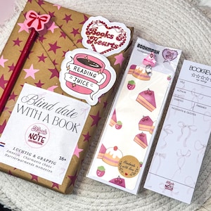 Puede incluir: Una colección de artículos con temática de libros, que incluye un libro envuelto con un lápiz de lazo rosa, pegatinas y marcadores. Las pegatinas dicen "Books & Hearts" y "Reading Juice". El marcador presenta rebanadas de pastel.
