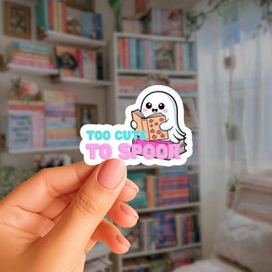 Könnte beinhalten: Ein weißer Aufkleber mit einem Cartoon-Geist, der ein Buch mit Kürbisillustrationen liest. Der Aufkleber hat den Text "Too Cute To Spook" in Blau und Rosa. Im Hintergrund ist ein Bücherregal mit Büchern zu sehen.