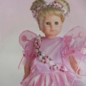 May include: A doll with blonde hair styled in two buns, wearing a pink dress with a floral sash and matching headband. The doll features translucent pink fairy wings with circular accents and a skirt with alternating light and dark pink stripes.