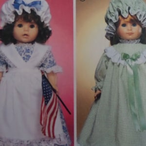May include: Two dolls dressed in vintage-style clothing. One doll wears a white apron over a blue floral dress and holds a small American flag. The other doll is in a green checkered dress with a matching bonnet and green ribbon.