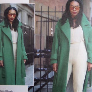 May include: A woman wearing a long, emerald green coat over a cream-colored outfit. The coat has a wide collar and a matching belt. The woman is also wearing orange-tinted sunglasses.