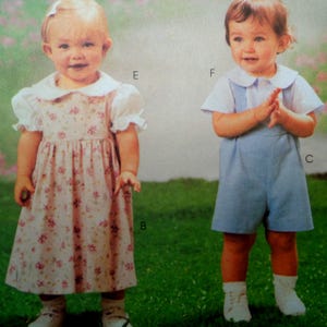 May include: Two children's outfits. One is a pink floral dress with a white collar and sleeves. The other is a light blue romper with a white collared shirt. Both children are wearing white socks and shoes, standing on green grass.