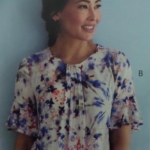 May include: A short-sleeved blouse featuring a white base with a watercolor-style print in shades of blue, purple, and red. The garment has a round neckline with a central vertical pleat detail and flared sleeves that extend to the elbows.