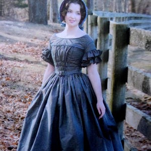 May include: A woman wearing a dark blue, checkered, vintage-style dress with short ruffled sleeves and a matching bonnet. The dress has a fitted bodice and a full skirt, typical of the era. The background shows a wooden fence and trees.