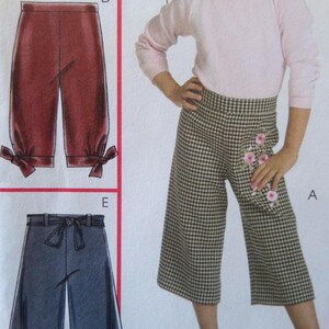 May include: A sewing pattern illustration featuring three styles of pants. Style A shows cropped pants with a black and white houndstooth pattern and pink floral embroidery. Style E displays dark blue pants with a tie waist and side bows.