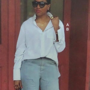 May include: A crisp white long-sleeved button-down shirt with a classic collar and cuffs, paired with light gray trousers. The shirt features a front pocket and is worn with a gold necklace and a gold ring. The letters A and B are visible on the right.