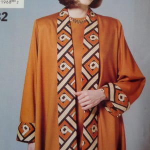May include: A solid burnt orange coat featuring long sleeves with wide cuffs and a front panel, both decorated with a geometric pattern in black, white, and orange. The coat is worn over a matching burnt orange top with a gold chain necklace.