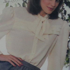 May include: A cream-colored long-sleeve blouse featuring a pussy-bow neckline, gathered shoulders, and a button-down front. The garment includes horizontal pleated detailing across the chest and is paired with a dark gray skirt.
