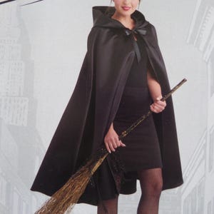 May include: A black hooded cape featuring a smooth, satin-like finish and a ribbon tie closure at the neck. The garment is worn over a black outfit and is paired with a long, rustic broomstick with natural brown bristles and a dark handle.
