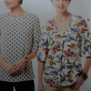 May include: Two women's tops with short sleeves. One top has a white background with a repeating geometric pattern in gray. The other top has a white background with a floral pattern in red, purple, and yellow. Both tops have a round neckline and a flared sleeve.