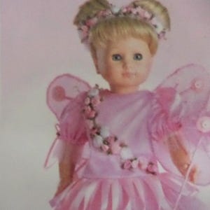 May include: A doll dressed as a fairy, with blonde hair and blue eyes. The doll wears a pink dress with white and pink accents, and sheer pink wings. The doll's hair is adorned with a floral headband.