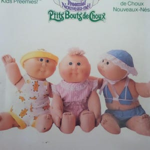 May include: Three Cabbage Patch Kids Preemies dolls with round faces and soft bodies. The left doll wears a yellow hat and white overalls with orange bear prints. The center doll wears a pink dress. The right doll wears a blue hat and blue outfit.