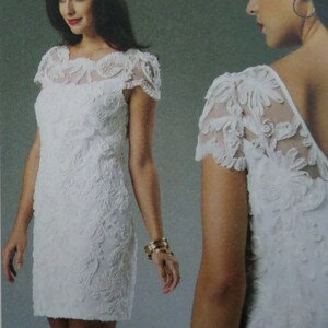 May include: A white knee-length dress featuring short sleeves and a sheer yoke with intricate floral embroidery. The garment displays a textured, raised pattern throughout the bodice and skirt, with a rounded neckline and a fitted silhouette.