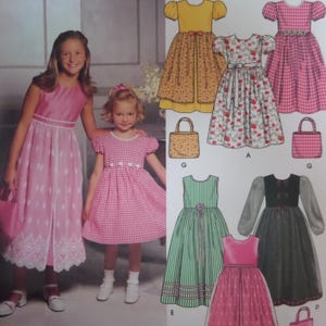 May include: A sewing pattern envelope featuring illustrations of various dresses and handbags. The designs include a yellow floral dress, a pink gingham dress, a white floral dress, a green striped dress, and a dark dress with sheer sleeves.