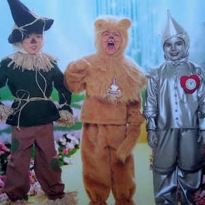 May include: Three children in costumes from The Wizard of Oz. The costumes include a scarecrow with a black hat and straw, a lion in a furry brown suit, and a tin man in a silver suit with a heart-shaped clock.
