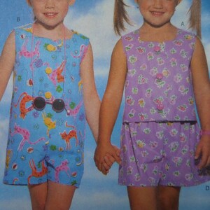 May include: Two children's outfits. One is a blue romper with a cat print, the other is a purple floral print top and shorts set. Both outfits have button closures. The children are holding hands.