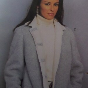 May include: A light gray jacket with a wide white lapel and a white turtleneck sweater underneath. The jacket features a button-down front closure with matching gray buttons. The letter D is visible on the right side of the image.
