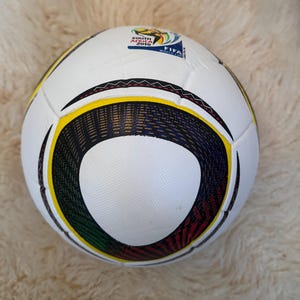 K&ouml;nnte beinhalten: Ein wei&szlig;er Fu&szlig;ball mit schwarzen, gelben, gr&uuml;nen und roten Akzenten. Der Ball hat ein kreisf&ouml;rmiges Design und den Text "SOUTH AFRICA 2010 FIFA WORLD CUP". Er liegt auf einer wei&szlig;en, flauschigen Oberfl&auml;che.