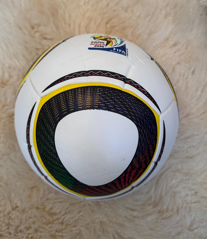 K&ouml;nnte beinhalten: Ein wei&szlig;er Fu&szlig;ball mit schwarzen, gelben, gr&uuml;nen und roten Akzenten. Der Ball hat ein kreisf&ouml;rmiges Design und den Text "SOUTH AFRICA 2010 FIFA WORLD CUP". Er liegt auf einer wei&szlig;en, flauschigen Oberfl&auml;che.