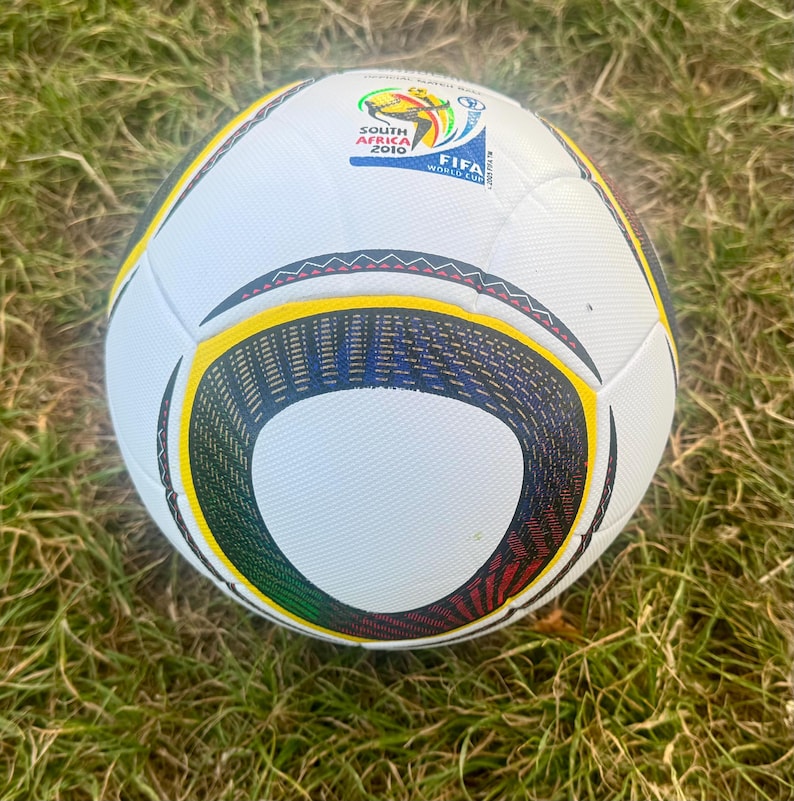 K&ouml;nnte beinhalten: Ein wei&szlig;er Fu&szlig;ball mit schwarzen, gelben, gr&uuml;nen und roten Akzenten. Der Ball hat ein Design mit geschwungenen schwarzen Linien und das offizielle FIFA World Cup South Africa 2010 Logo.