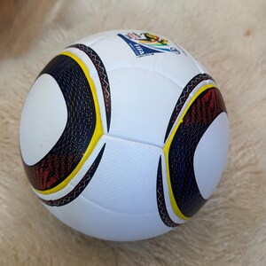 K&ouml;nnte beinhalten: Wei&szlig;er Fu&szlig;ball mit schwarzen, gelben und roten geschwungenen Akzenten. Mit dem Logo der FIFA Fussball-Weltmeisterschaft 2006. Der Ball hat eine strukturierte Oberfl&auml;che.